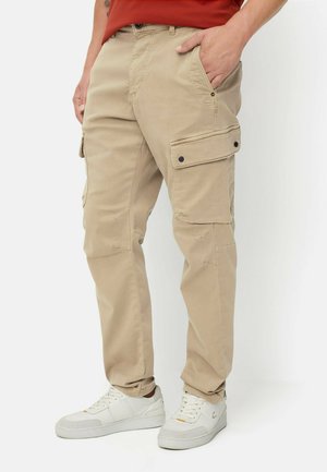 Beige Cargohose mit seitlichen Klappentaschen, getragen mit weißen Sneakern und einem roten Hemd, gezeigt von der Taille bis zu den Füßen.
