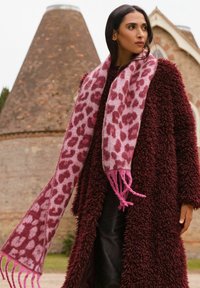 Bufanda de estampado de leopardo en tonos rosa y burdeos, hecha de un tejido suave con bordes flecos. Llevada sobre un abrigo texturizado de un profundo color burdeos.