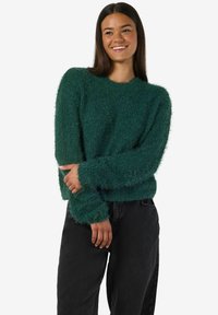 Zelena dlačasta pulover s dolgimi rokavi, rebrastimi manšeti in robom, ki ima mehak značaj in sproščen kroj, v kombinaciji s črnimi hlačami.