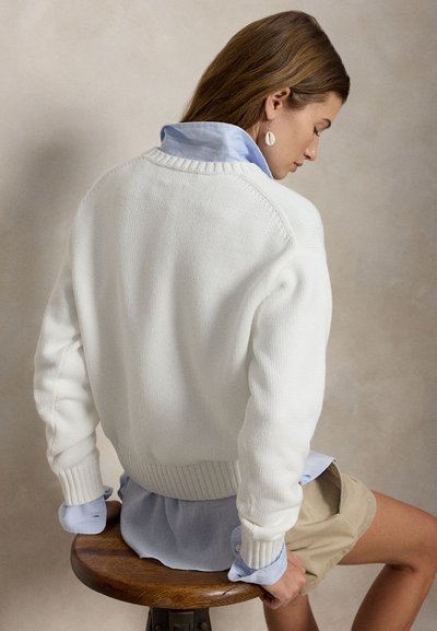 Νέα γυναίκα με ανοιχτό καστανά μαλλιά, φορώντας λευκό πλεκτό πουλόβερ, ανοιχτό μπλε πουκάμισο, μπεζ σορτς και σκουλαρίκια με κοχύλια, καθισμένη σε ένα ξύλινο σκαμπό.