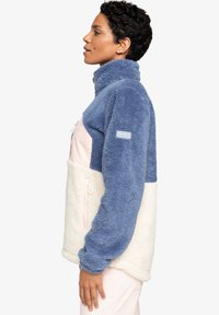 Fleece-pullover i blå og krem, med høy krage, myk tekstur, sidelommer og en rosa brystpanel. Har et "ROXY" logo-patch.