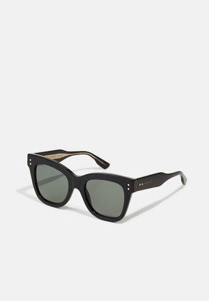 Gafas de sol negras en forma de cuadrado con lentes verdes, montura de acetato gruesa y acentos dorados. Detalles notables incluyen pequeños remaches en los laterales.