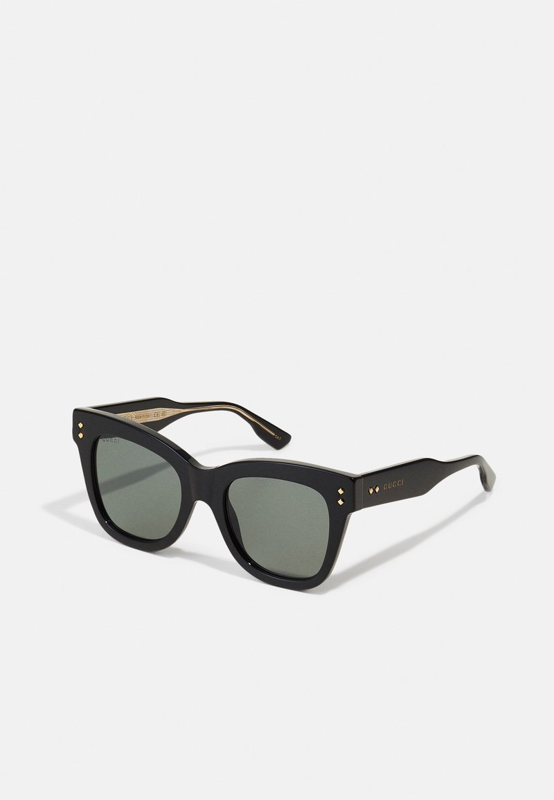 Melni kvadrātveida saulesbrilles ar zaļiem stikliem, biezām acetāta rāmēm un zeltītiem akcentiem. Pamanāmas detaļas ietver mazus pērlītes sānos.