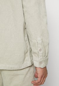 Manga de camisa de pana beige claro con puño abotonado y pantalones a juego, mano relajada al lado contra un fondo liso.