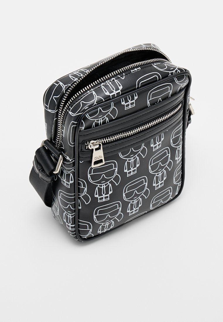 Bolso cruzado negro hecho de material sintético texturizado, con personajes de dibujos animados blancos con gafas de sol. Compartimentos de doble cremallera con herrajes plateados.