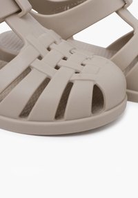 Chanclas de plástico de color beige claro con un diseño de tira trenzada, puntera circular y plantilla texturizada para mayor agarre. Lados abiertos y suela resistente.