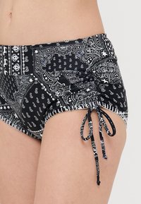 Plavky s černobílým vzorem, s elastickým pasem, bočním řasením a nastavitelnými šňůrkami na spodním lemu. Vyrobeno z měkkého a pružného materiálu.