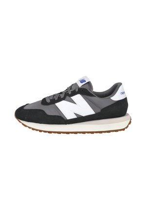Zapatilla New Balance gris y negra con logo "N" blanco, lengüeta trasera blanca, cordones grises y suela de goma marrón.