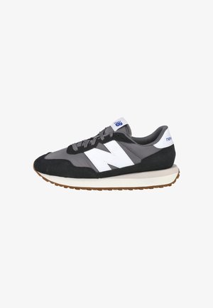 Zapatilla New Balance gris y negra con logo "N" blanco, lengüeta trasera blanca, cordones grises y suela de goma marrón.
