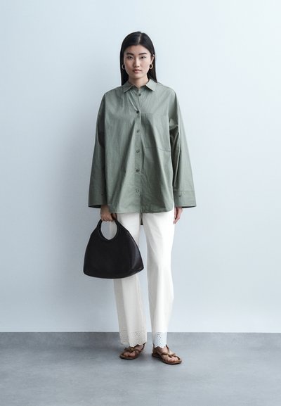 Νεαρή γυναίκα που φοράει υπερμεγέθη πράσινο πουκάμισο, λευκό παντελόνι με τελείωμα από δαντέλα, καφέ σανδάλια, κρατώντας μαύρη τσάντα χειρός σε απλό φωτεινό φόντο.