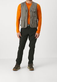 Harmaa hyötyliivi, jossa on useita taskuja oranssin pitkähihasen paidan päällä, yhdistetty mustiin housuihin ja ruskeisiin vaelluskengiin.