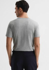 Tricou gri cu mânecă scurtă, realizat dintr-un material moale, având un design clasic cu gât rotund, purtat împreună cu pantaloni navy închisi, văzut din spate.