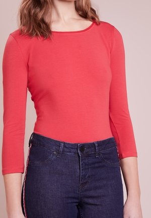 Femme portant un haut ajusté à manches longues corail et un jean taille haute bleu foncé avec une bande latérale rouge et blanche.