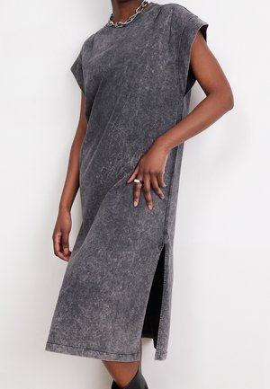 Osoba v bezrukávových vybledlých černých midi šatech s bočním rozparkem, stříbrném řetízkovém náhrdelníku a stříbrném prstenu na levé ruce.