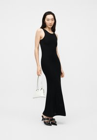 Γυναίκα με μαύρο πλεκτό μακρύ φόρεμα χωρίς μανίκια, μαύρα ψηλοτάκουνα παπούτσια με λουράκια και μυτερό σχέδιο, κρατώντας μια λευκή τσάντα χειρός μπροστά σε απλό φόντο.