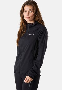 Woman with long brown hair wearing a black zip-up hooded jacket and black leggings with brand logos, standing against a plain background.