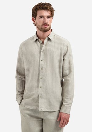 Mann trägt ein hellbeiges, längsgestreiftes Hemd mit langen Ärmeln und passenden Hosen, steht mit einer Hand in der Tasche.