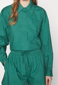 Camisa verde de mangas largas y cuello puntiagudo, adornada con lentejuelas reflectantes. Combinada con pantalones cortos de cintura elástica a juego.