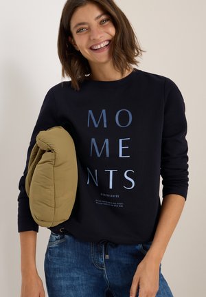 Sweatshirt bleu marine avec le texte "MOMENTS" en bleu clair, associé à un coussin beige. Il présente un col rond et un ourlet à cordon de serrage.