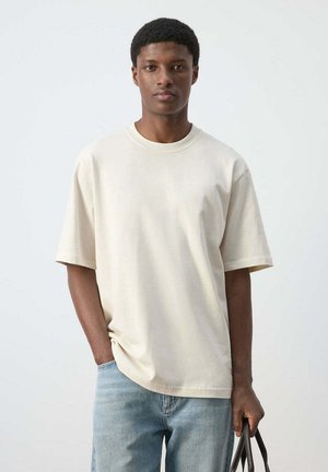 Jeune homme portant un t-shirt oversize écru et un jean bleu clair, tenant la poignée d’un sac marron foncé sur un fond neutre.