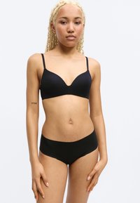 Νεαρή γυναίκα με ανοιχτόξανθες πλεξούδες, φορώντας μαύρο σουτιέν και μαύρο εσώρουχο, στέκεται μπροστά σε ένα απλό ανοιχτόχρωμο φόντο.