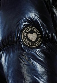 Navy blue puffy jacket with glossy texture, featuring a circular black patch with embroidered heart and text "Be Happy Killed".