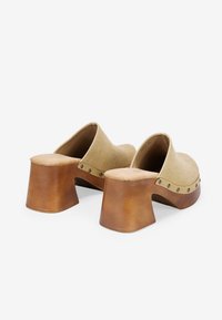 Zuecos de ante beige con un ancho tacón de madera marrón y tachuelas en tono plateado a lo largo del borde superior. Textura suave y diseño minimalista.