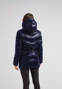 Marineblå pufferjakke med en skinnende finish, indsnævret taljedetalje og en stor hætte. Har chevron-syninger og elastiske manchetter.