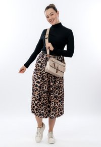 Lächelnde Frau mit einem schwarzen langärmligen Oberteil, einem Rock mit Leopardenmuster, weißen Sneakers und einer beigen Umhängetasche vor einem schlichten weißen Hintergrund.