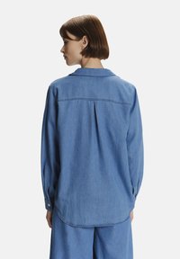 Camicia in denim blu con colletto, maniche lunghe e un piegone sul retro per una forma extra. Tessuto morbido e polsini con bottoni.