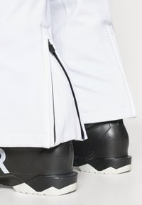 Fehér sínadrág oldalán cipzárral, fekete csizmával párosítva, melyen texturált felület és a talpon fehér cikk-cakk minta található.