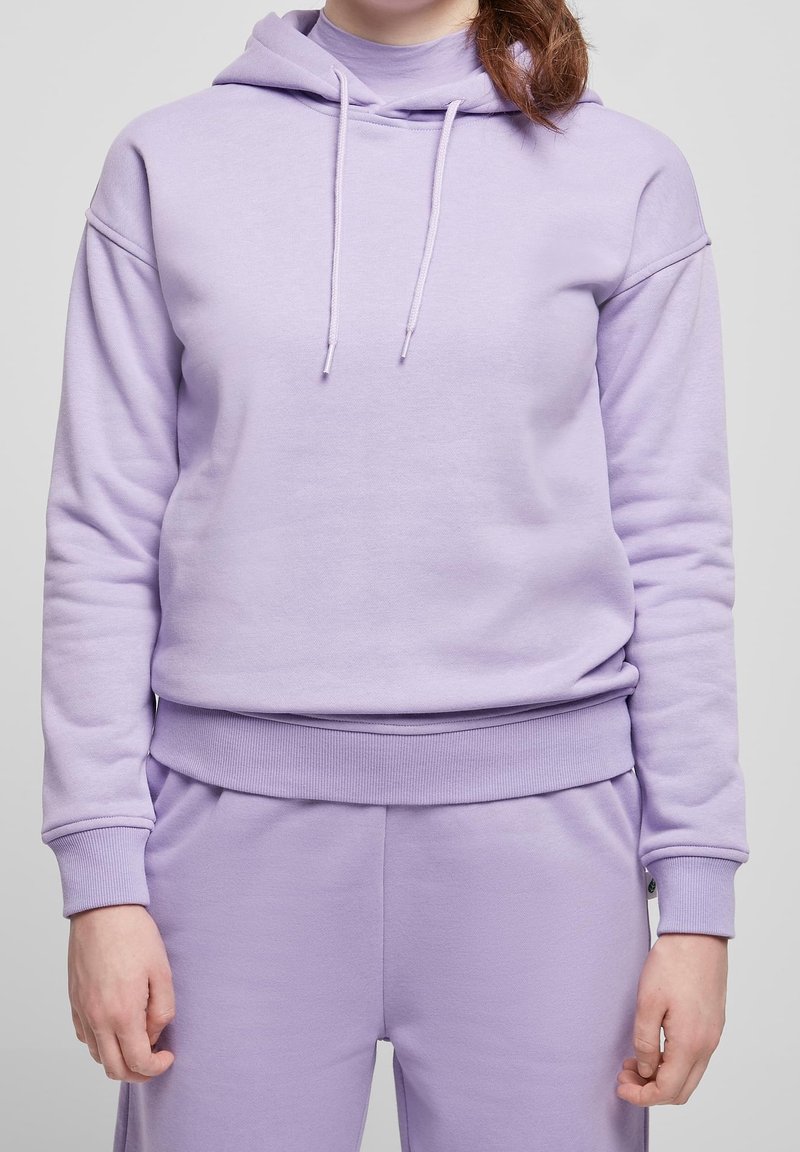 Personne portant un sweat à capuche violet clair et un pantalon assorti, avec les cheveux attachés en queue de cheval basse, debout devant un fond uni.