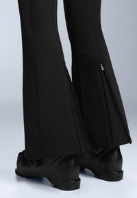 Fekete, harangszabású nadrág, cipzáras mandzsettákkal, sima, rugalmas anyagból készült. Masszív fekete cipővel viselve, amely texturált talppal rendelkezik.