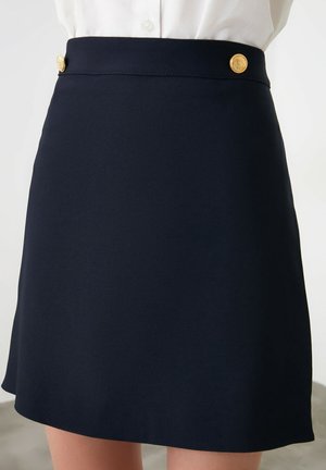 Jupe trapèze bleu marine à taille haute, décorée de deux boutons dorés, portée avec une chemise blanche rentrée.