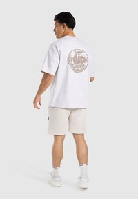 Camiseta de manga corta gris claro con un gráfico circular en la parte posterior. Combinada con pantalones cortos gris claro y zapatillas blancas, todo ello sobre un fondo neutro.