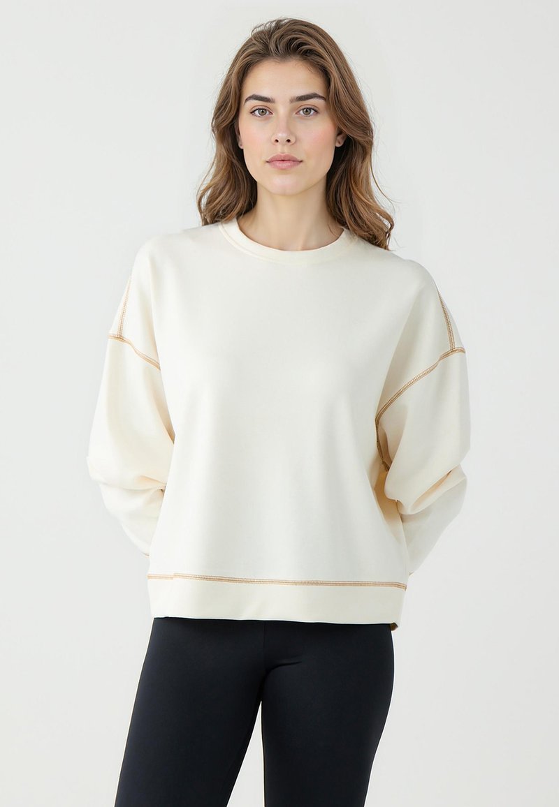 Sweatshirt couleur crème avec des manches ballon, couture beige contrastante le long des coutures et un col rond classique. À associer avec des leggings noirs.