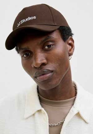 Bruine baseballpet met witte borduring van "JJ Studios", gedragen door een persoon met middel-lang donker haar, in laagjes met een textuur van een witte shirt.
