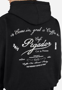 Black hoodie featuring white text graphics, including "Café Pigador" and coffee-related phrases, with a drawstring hood and soft texture.