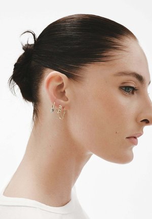 Pendientes circulares de oro y plata en la oreja de un modelo; el diseño moderno presenta texturas y formas mixtas, exhibiéndose sobre un fondo neutro.