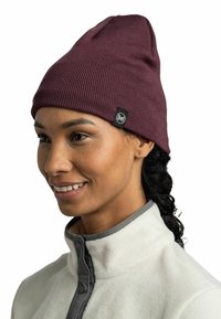 Burgundy knit beanie with a tapered design, ribbed texture, and a black Buff logo tag. Worn with a light fleece jacket.