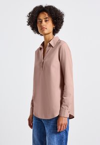 Blusa a maniche lunghe rosa chiaro con colletto classico, apertura con un solo bottone e orlo arrotondato. Texture morbida e liscia. Abbinata a jeans blu.