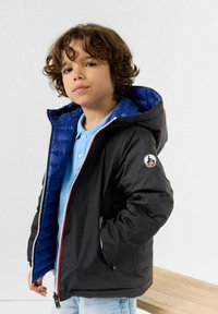 Giacca a vento nera con fodera trapuntata blu, dettagli con zip rossi e un patch con logo sulla manica. Indossata sopra una maglietta azzurra chiara.