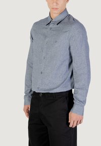Camicia abbottonata in tessuto leggero blu chiaro con colletto, maniche lunghe e bottoni scuri, abbinata a pantaloni neri.