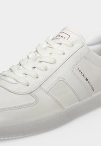 Λευκό sneaker χαμηλού τύπου με λευκά κορδόνια και το λογότυπο Tommy Hilfiger στο πλάι και τη γλώσσα πάνω σε λευκό φόντο.
