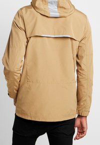 Giacca impermeabile color sabbia con cappuccio, dotata di una ventilazione posteriore con zip, polsini elastici e dettagli riflettenti. Texture liscia, vestibilità casual.
