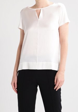 Vrouw draagt een witte blouse met korte mouwen en een keyhole-halslijn en zwarte broek tegen een effen achtergrond.