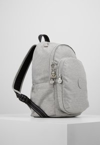 Šedý látkový batoh s kulatým tvarem, se zapínacími přihrádkami, gumovým logem a černým pruhovaným nastavitelným popruhem.