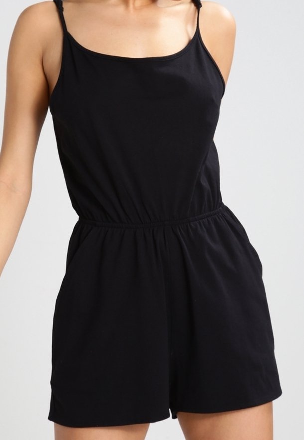 Femme portant une combinaison noire sans manches avec des bretelles fines et une taille élastique, debout devant un fond clair uni.