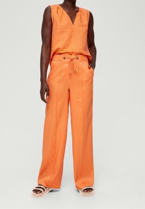 Personne portant un haut orange sans manches et un pantalon large assorti avec cordon de serrage, associé à des sandales légères, debout devant un fond uni.