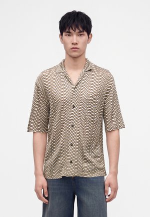 Junger Mann mit kurzen schwarzen Haaren, der ein kurzärmeliges, gemustertes Hemd mit Knöpfen und blaue Jeans trägt, steht vor einem schlichten Hintergrund.